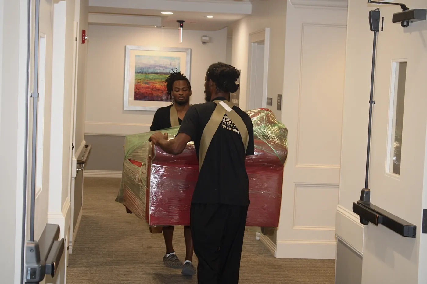 Two GoldStrap Moving crew members carrying a wrapped sofa down a hallway with shoulder straps