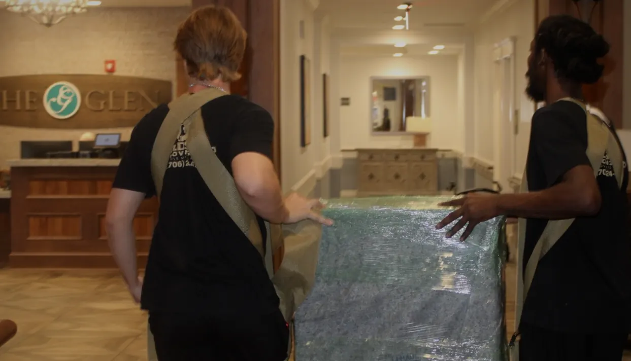 Two GoldStrap Moving crew members carrying blanket-padded furniture through a lobby near Lake Oconee