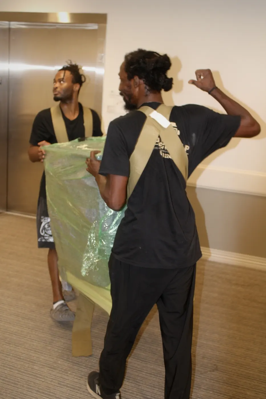 Two GoldStrap Moving crew members carrying wrapped furniture out of an elevator lobby