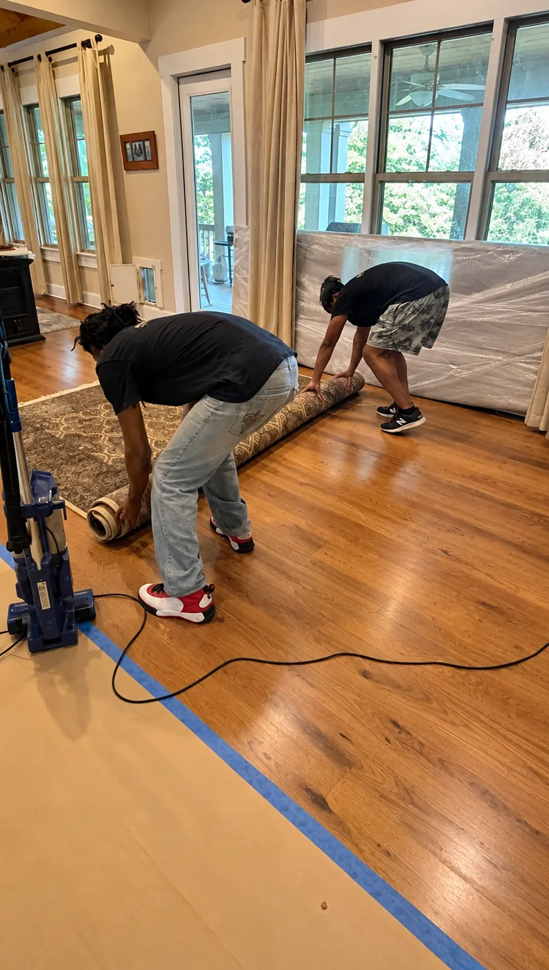 Two movers rolling a rug in a living room, with a wrapped mattress against the windows and floor protection laid down