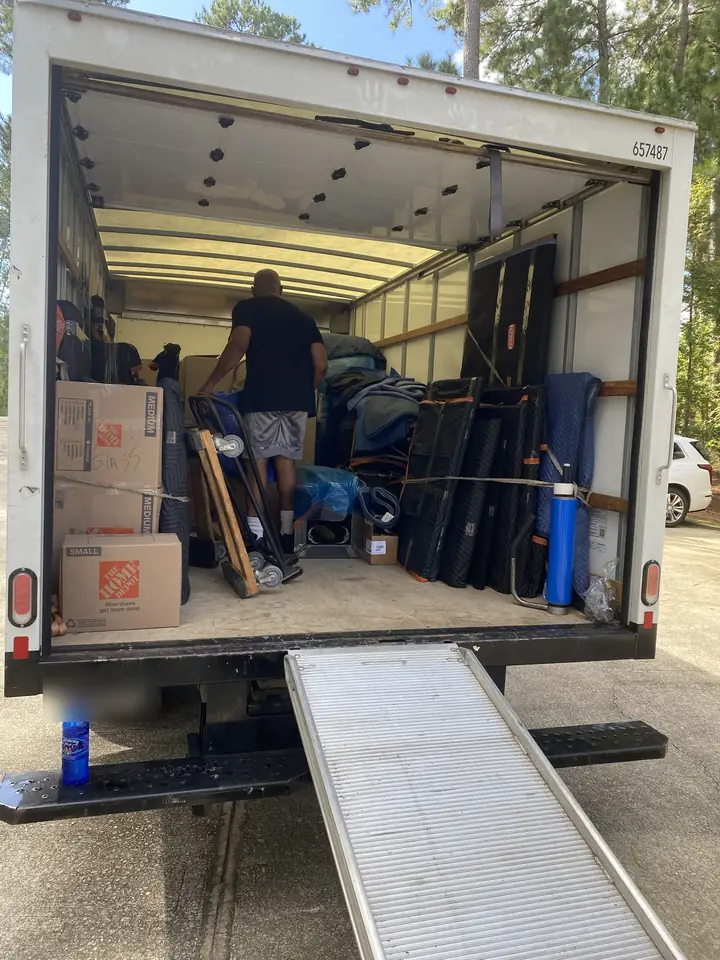 Inside a moving truck with furniture wrapped in moving blankets, a hand truck, stretch wrap and boxes, and a loading ramp
